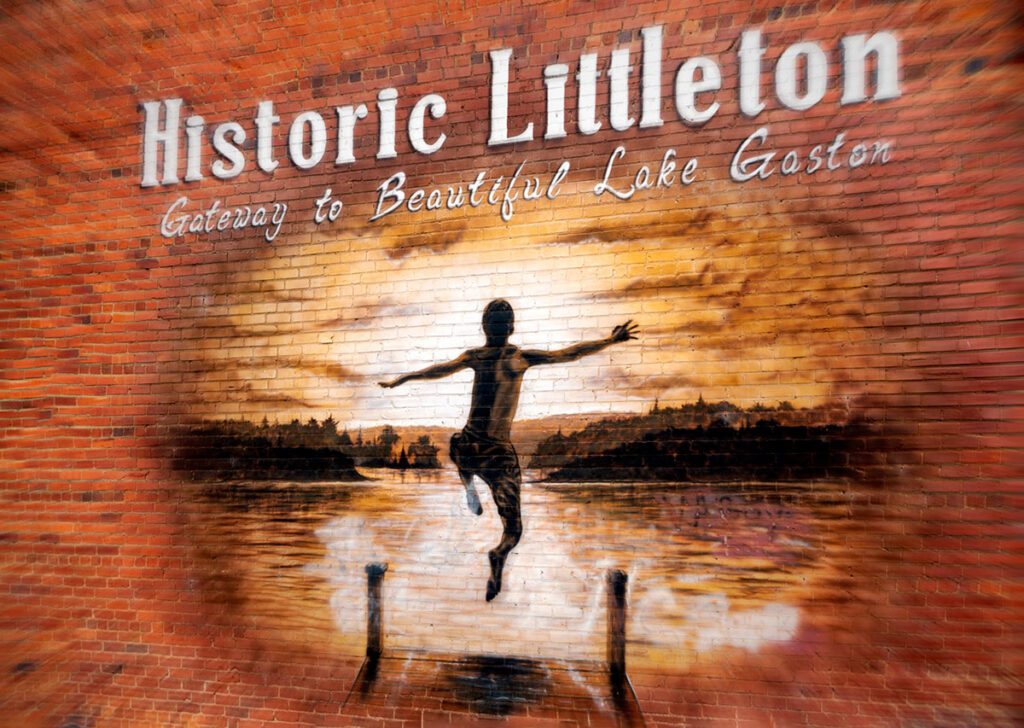 A beautiful mural painted on a brick wall of a boy jumping off the end of a dock into a lake at sunset. The words, "Historic Littleton, Gateway to Beautiful Lake Gaston" run across the top of the painting 