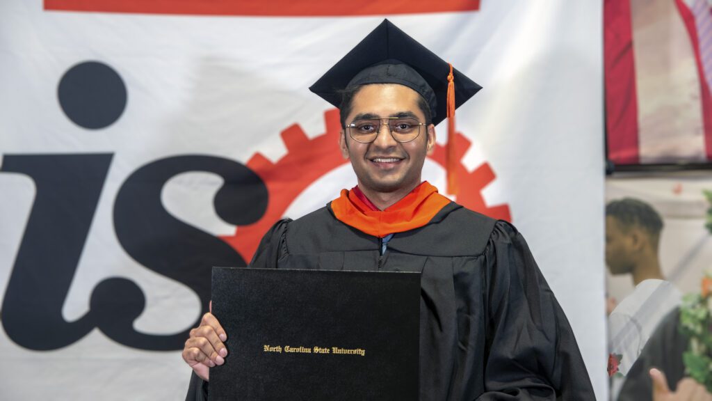 MEM student Kartik Khanderia holds up his diploma in front of a big ISE backdrop