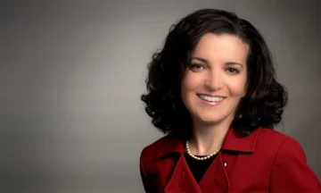 A headshot of Elif Oker standing in front of a medium gray background.