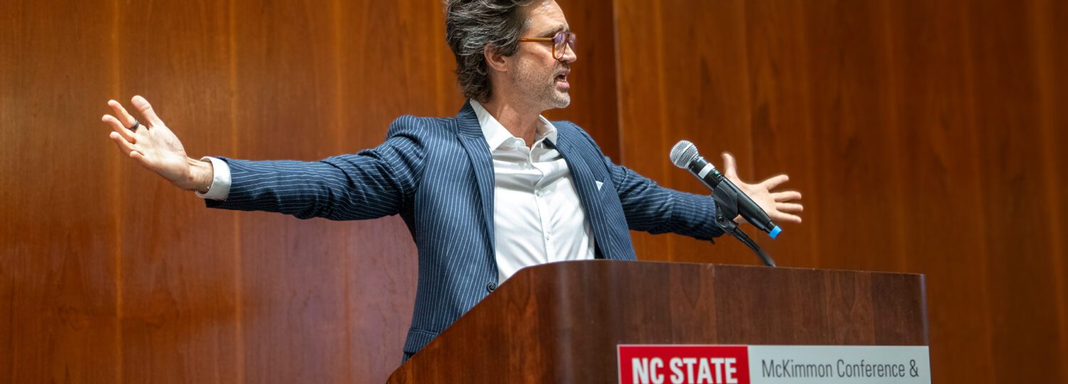 Link Neal standing behind the podium with his arms spread out wide.