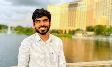 Dwij Bharadwaj stands on a bridge in front of a lake and a hotel