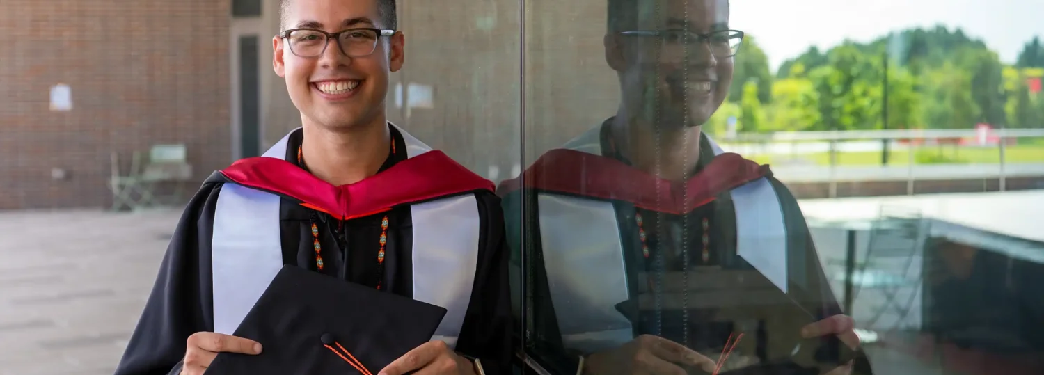 Michael Sanderson in his graduation cap and gown standing next to his reflection in the glass windows of Fitts-Woolard Hall