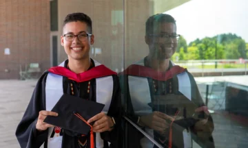 Michael Sanderson in his graduation cap and gown standing next to his reflection in the glass windows of Fitts-Woolard Hall