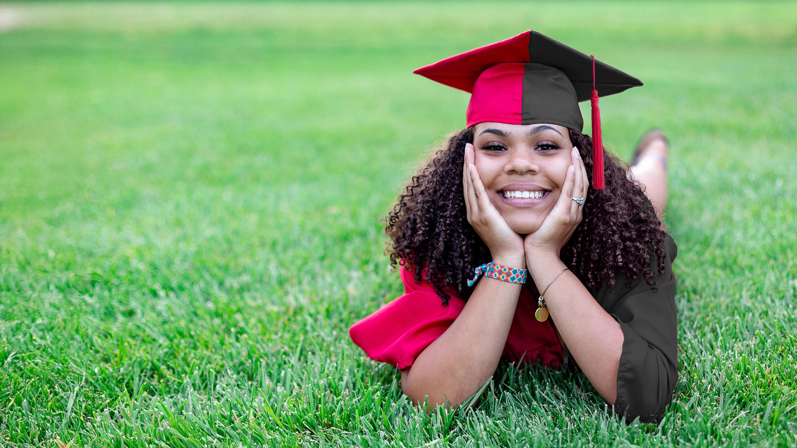 A female student laying on the grass in a two tone graduation cap and gown. The two colors represent degrees of the Accelerated Bachelors/Masters Program.