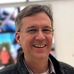 A headshot of ROb Drew standing in front of a wall of screens.
