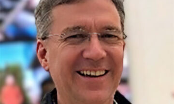 A headshot of ROb Drew standing in front of a wall of screens.